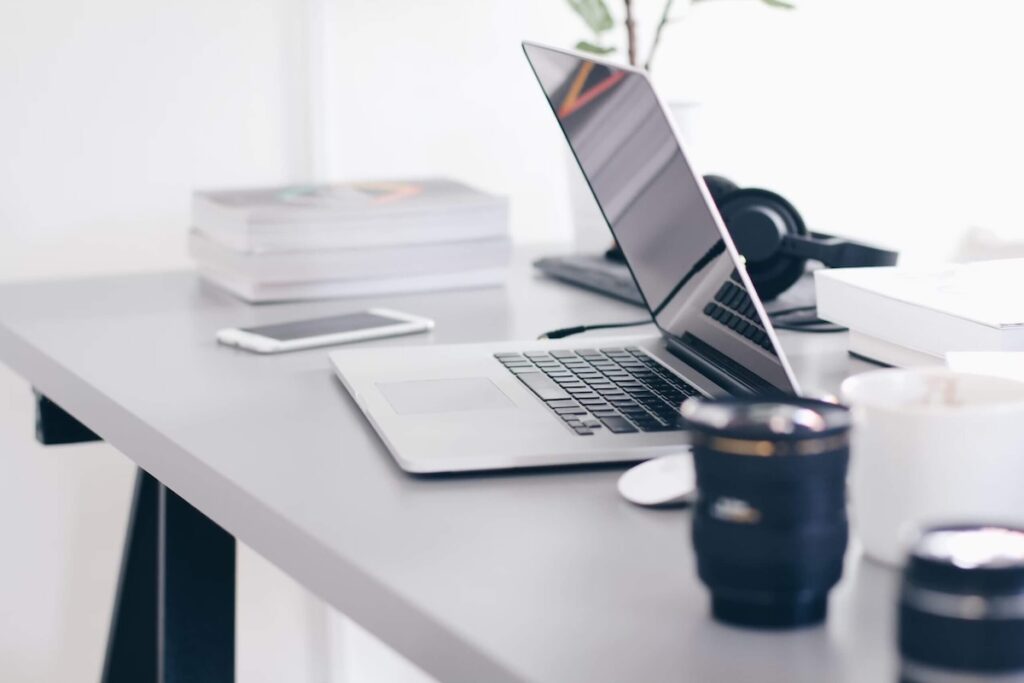 Macbook laptop stående på skrivbord. Ur fokus finns mobil, hörlurar, kamera linser, kaffekopp och böcker.